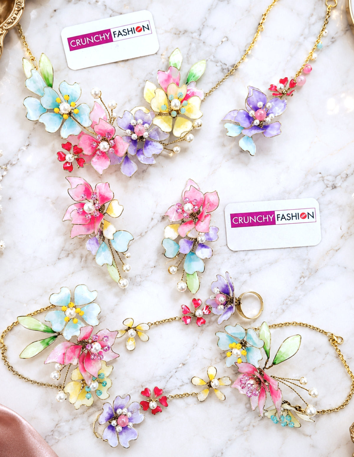 A flat-lay display of jewelry from the brand Crunchy Fashion, featuring floral necklaces and matching earrings with colorful enamel flowers in pink, blue, purple, yellow, and green on gold chains, set against a white marble background.